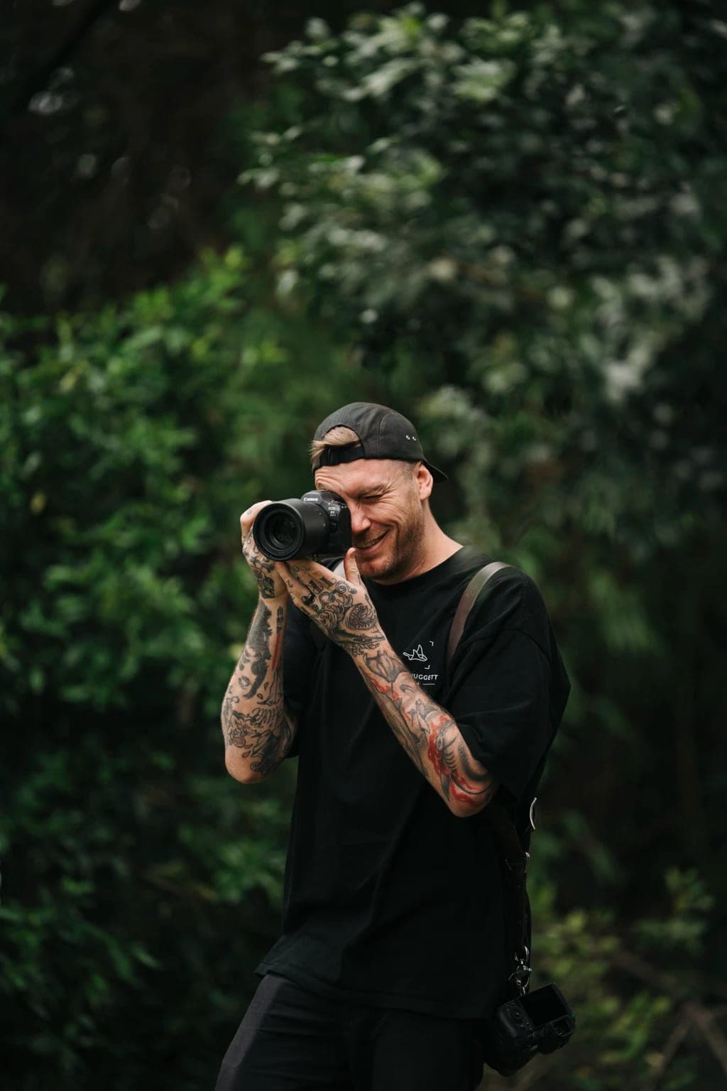 Josh Huggett, in a cap and tattoos, laughing behind his camera among green trees
