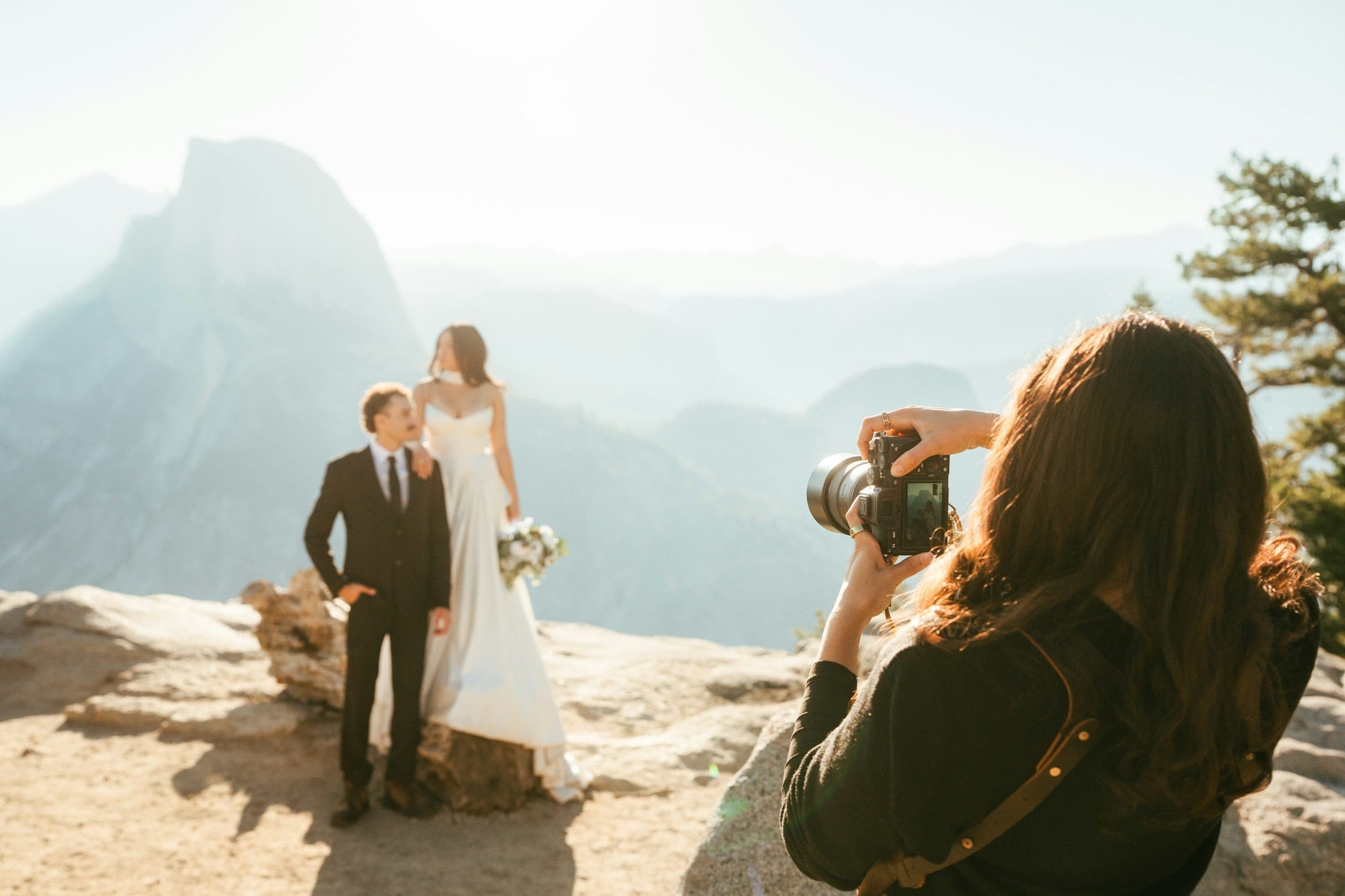 A wedding photographer framing a bride and groom on a mountain lookout in soft morning light